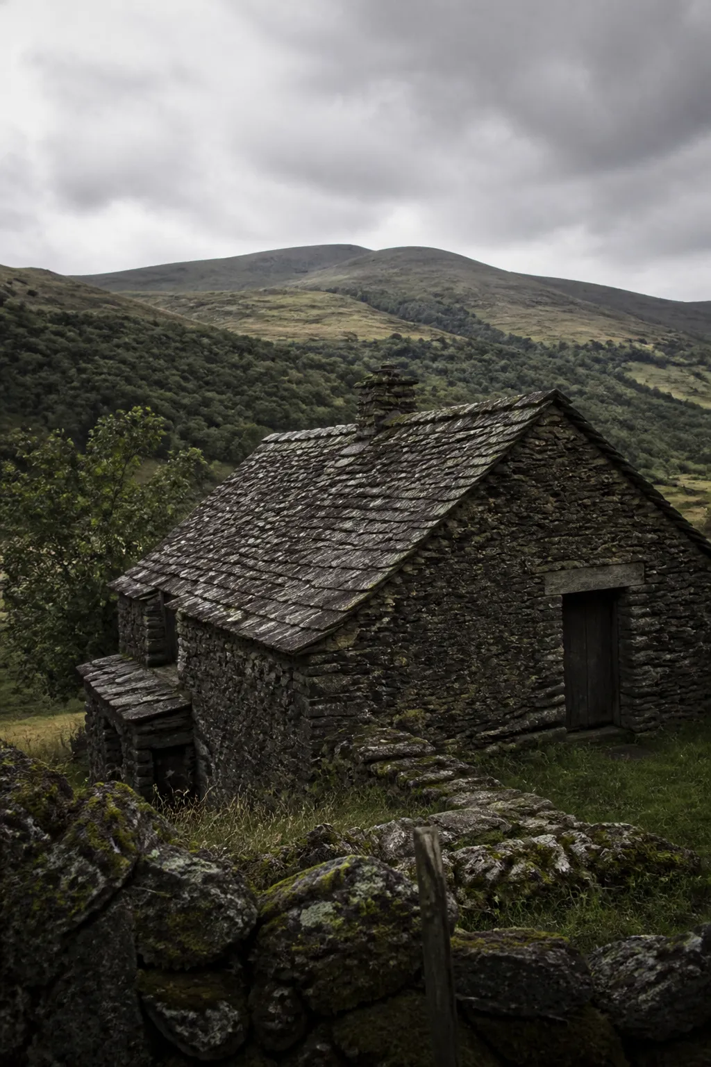 Maison en pierre sombre avec toiture en lauzes dans un paysage de moyenne montagne