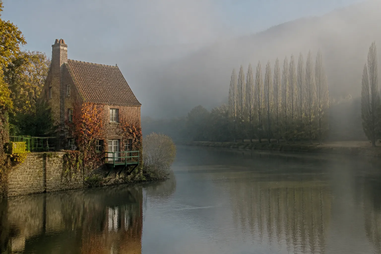 Maison au bord d’une rivière bordée d’arbres en fond de vallée